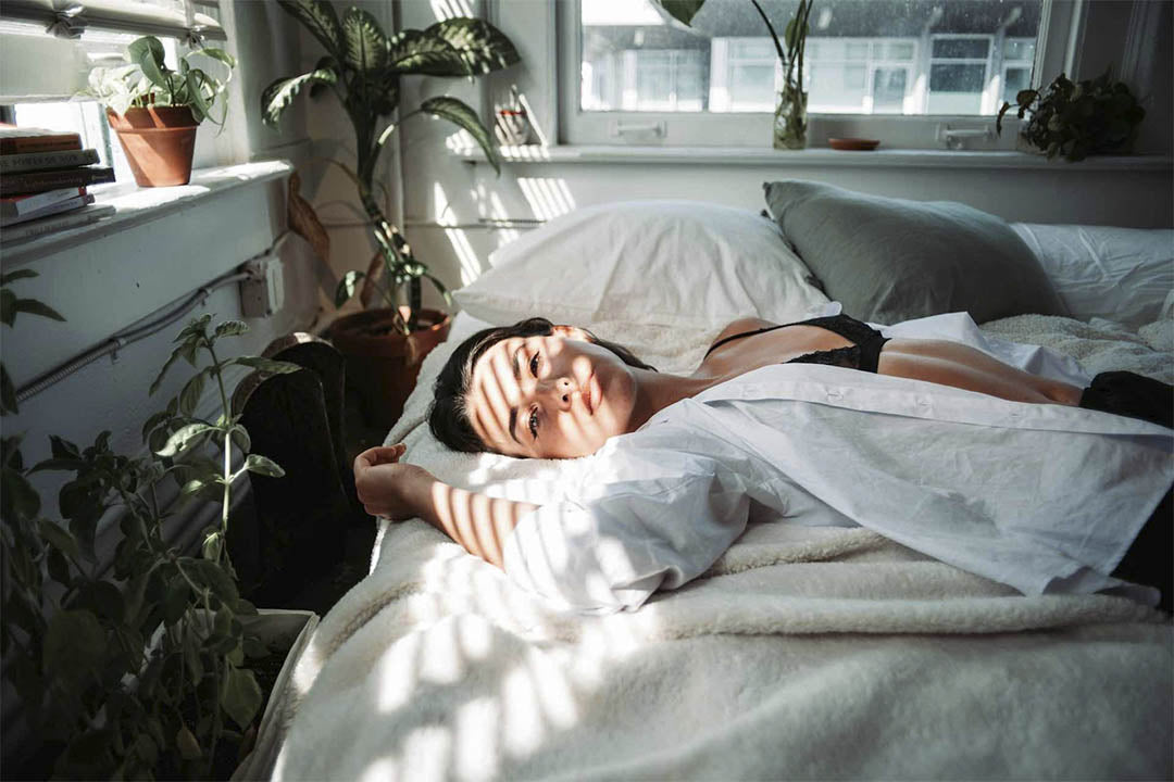 Sinnliche Frühlingsgefühle: Frau in schwarzen Dessous und weißer Bluse im warmen Sonnenlicht auf dem Bett – Magische Lingerie-Momente.
